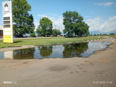 A digital photograph captures a reflection of trees and sky in a large puddle on an asphalt surface. To the left, a gas station price board displays "РОСНЕФТЬ" and fuel prices of 92, 95, and AT, with numerical values of 53.00, 59.40, and 56.10 respectively. In the background, lush green trees line a grassy area, and several cows graze under a clear blue sky with scattered white clouds. The timestamp "16.07.2026 11:40" and the text "REDMI 13C" are visible in the bottom right and left corners, respectively. The scene appears to be located at a gas station on the outskirts of Nikol'sk, Russia, judging by the "РОСНЕФТЬ" signage which is a prominent Russian oil company.