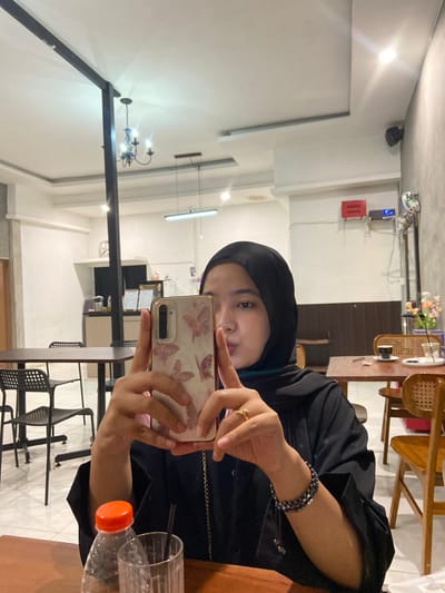In this image, a young woman wearing a black hijab and a black outer garment is taking a selfie in a dimly lit indoor setting. She is holding up a smartphone with a delicate floral pattern on its case, its camera facing the mirror to capture her reflection. A beaded bracelet adorns her left wrist, and a delicate ring is visible on her index finger.
The background suggests a cafe or restaurant environment. In the upper left, a black security camera is mounted on the ceiling. Further back, a red fire alarm box is visible on a white wall, next to a blue control panel. The decor includes wooden chairs and tables, and a chandelier with multiple lights hangs from the ceiling. A glass of water with a straw and a partially filled plastic bottle with an orange cap are on a table in the foreground.
The lighting is somewhat stark, with bright spots from overhead lights casting shadows. The woman's expression is neutral as she focuses on her phone. The overall scene appears to be a casual moment captured within an establishment in Sekar Mawar, Indonesia. The time of day is not clearly indicated, but the indoor setting suggests it could be evening or a time with less natural light.