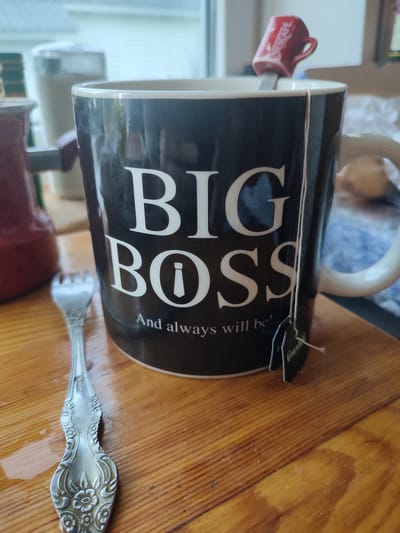 A close-up, eye-level shot captures a black mug with "BIG BOSS" printed in large white letters, followed by "And always will be!" in smaller white font. A small, red Nescafé-branded spoon rests across the mug's rim. A green tea bag tag hangs over the side of the mug. To the left of the mug, a silver fork with an ornate floral pattern rests on a wooden surface. In the background, out of focus, is a kitchen setting with a window and other kitchen appliances. The scene is set on a wooden table. The image does not provide any information about the city of Velikii Krai, Russia.