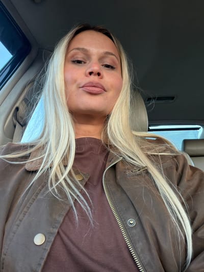 A young woman with long, blonde hair is sitting in a vehicle. She is wearing a brown jacket over a brown top. The interior of the vehicle is visible, including the dashboard, window, and seat. There is no indication she is in Дулесово, Russia.