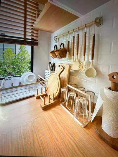 The image depicts a well-organized kitchen counter area with a focus on kitchenware and storage. In the background, a window with horizontal wooden blinds offers a view of lush green trees. A white subway tile backsplash covers the wall behind the counter.
Mounted on the wall is a wooden rod with several kitchen utensils hanging from S-hooks. These utensils have wooden handles and include various spatulas and a pasta server. Two small woven baskets are also hanging from the rod. To the left of the utensils, a white rack holds several knives.
Dominating the center of the image is a white dish rack containing plates and utensils, positioned near a sink area. Adjacent to the dish rack, a wooden cutting board with a distinctive curved handle stands upright. In front of the cutting board, a wooden rack holds several clear glasses, some upright and some inverted.
To the right of the glasses, a roll of paper towels is mounted on a wooden dispenser topped with a decorative wooden flower. The countertop itself is made of light-colored wood with a prominent grain pattern. The overall impression is one of a clean, functional, and aesthetically pleasing kitchen space. The image does not contain any elements that would allow for identification of its specific location in Matangpanyang, Indonesia.