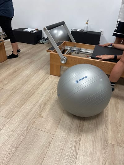 A gray exercise ball sits on a light-colored, wood-grain floor. Adjacent to the ball is a Pilates reformer machine, with a light-colored wood frame and black platform. Parts of two people are visible, including hands on the reformer's platform and feet near the machine. A black cabinet with white paper on top, and a water bottle are located near the reformer.