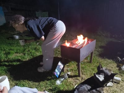 The image depicts a nighttime scene outdoors, likely in a rural area such as Патоцкі сельскі Савет, Belarus. In the foreground, a person is bending over a small metal barbecue grill with a fire burning inside, holding a white mug. The person is wearing a dark t-shirt and light-colored pants, with white clogs on their feet. Beside the grill, a tablet computer is propped up, displaying content. A clear plastic bottle lies on the grass nearby. In the lower right portion of the image, a dog, appearing to be a husky mix, is lying down. The environment consists of green grass and dark, indistinct background elements suggesting it is nighttime.