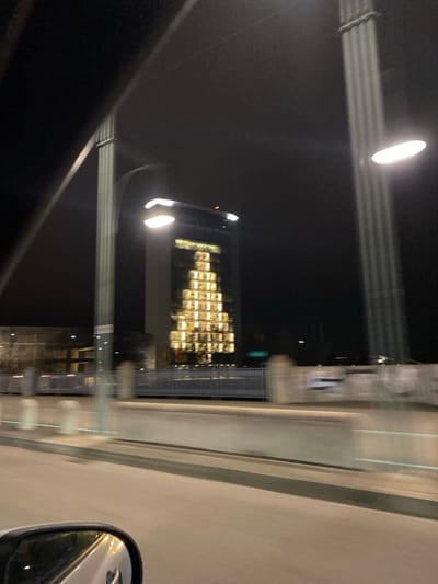Nighttime view from a car, capturing a building with windows lit to resemble a Christmas tree. Streetlights and road infrastructure are blurred in the foreground. The car's side mirror is visible on the right edge. The overall scene has a dark, blurry quality, suggesting movement and low light conditions.