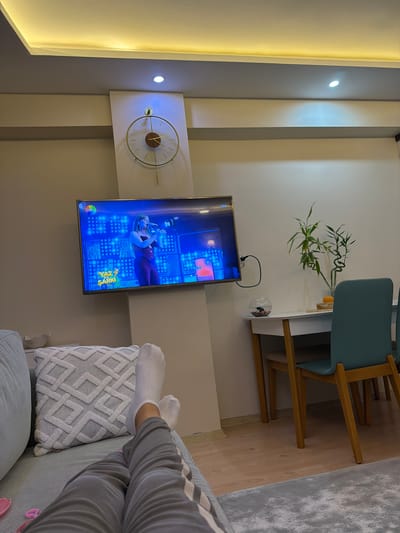 The image shows a living room, likely in Manisa, Turkey, with a television displaying a musical performance. A person's legs, clad in grey sweatpants and white socks, are propped up on a grey couch in the foreground, facing the television. To the right of the television, a white dining table with two teal chairs is visible. On the table, there is a vase with a bamboo plant and a small fishbowl containing a black and white fish. A modern clock with a gold frame hangs on the wall above the television. The room has a neutral color scheme with beige walls and a grey rug. The lighting appears to be from recessed ceiling lights and ambient light from the television.