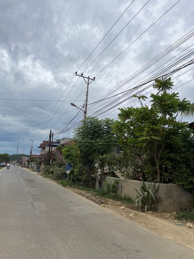 The image depicts a street scene in Rantau Pauh, Indonesia, under a cloudy sky. An asphalt road runs from the foreground towards the left, with a few potholes visible. On the right side of the road, a concrete wall borders a property with lush green vegetation and a large tree with compound leaves. Several utility poles with numerous electrical wires crisscross overhead. Along the left side of the road, houses and buildings with varying rooflines are visible, interspersed with more trees and foliage. A few motorcycles and people are present on the road in the distance, indicating ongoing activity. The overall environment suggests a semi-urban or rural setting with a mix of infrastructure and natural elements.