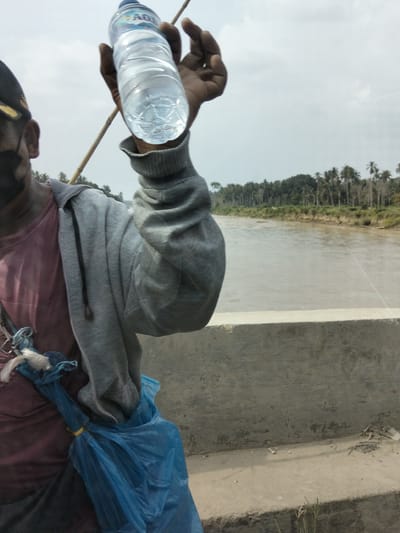A person is holding a bottle of water in their right hand, with their arm extended upwards. They are wearing a maroon shirt and a grey hooded jacket. A blue plastic bag is tied to their left side with a yellow band. The person is standing on a concrete structure, possibly a bridge or embankment, overlooking a wide, muddy river. The river is bordered by a dense line of palm trees and other vegetation. The sky is overcast, suggesting a cloudy day. The time of day appears to be daylight. The person is wearing a dark cap and a black face mask. There is a bamboo pole or stick held by the person's left hand. The brand "AQUA" is visible on the water bottle. The location is Teupinreudeup, Indonesia.