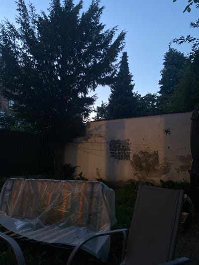 The image captures a dimly lit backyard scene, likely in Hildesheim, Germany, given the context. The foreground shows two outdoor chairs, one with a textured dark gray seat and back, partially obscuring a bench covered in translucent plastic sheeting. The bench is striped and appears to be made of metal. In the mid-ground, a weathered white wall runs horizontally across the frame. The wall exhibits cracks and discolored patches, with what appears to be a grid of glass blocks set into it, forming a window-like feature. Several clotheslines with clothespins are stretched across the wall, though no clothes are visible. To the left of the wall, a dark, tall fence can be seen. The background is dominated by the silhouettes of various trees against a clear blue twilight sky. A large evergreen tree with dense foliage stands prominently on the left, while other coniferous trees of varying heights are visible behind the wall and fence. The overall impression is of a quiet, residential backyard at dusk.