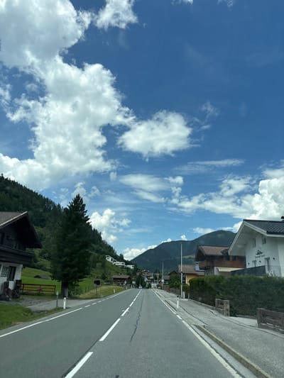The image depicts a scenic view of a road in Bruck an der Großglocknerstraße, Austria, on a bright, partly cloudy day. The road, paved with asphalt and marked with white lines, curves gently into the distance, flanked by lush green hillsides on the left and a line of buildings and dense hedges on the right.
Dominating the left side of the frame is a steep, tree-covered mountain slope, with the dark-roofed facade of a building visible at the bottom left. A tall, dark green evergreen tree stands prominently near the road. On the right, several houses with traditional alpine architecture, featuring tiled roofs and balconies, are nestled behind manicured hedges. One white building has a logo depicting a mountain and a bird, with the text "Apartments Kolb" below it.
The sky is a vibrant blue, adorned with scattered white cumulus clouds, suggesting fair weather. The overall impression is one of a peaceful rural setting, typical of the Austrian Alps. There are no people or animals visible in the image. The road appears to be a well-maintained public thoroughfare.