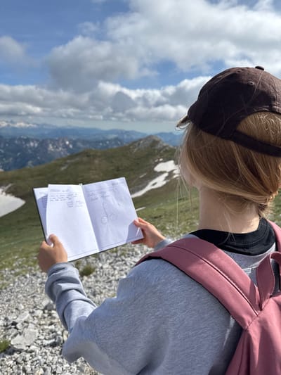 A person with blonde hair and a brown baseball cap is standing on a rocky mountain slope, holding open a notebook. The notebook contains handwritten text and a drawing of a penis. The person is wearing a grey sweatshirt and a pink backpack. In the background, there are more mountains and a cloudy sky. The location is Jošanica, Montenegro.