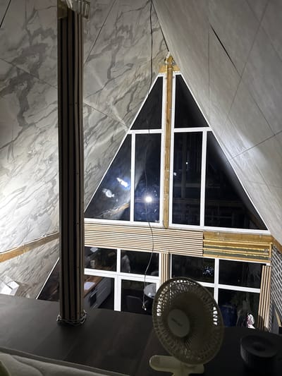 An interior shot captures a portion of a house with a triangular window, suggesting an A-frame or modern architectural style. The walls are covered in large marble-patterned tiles. A wooden column, wrapped with a metal chain at its base, stands prominently in the foreground. To the right of the column, a white oscillating fan sits on a dark surface. The triangular window reveals a dark interior with some reflections, hinting at the outside being dark. Below the triangular window, there is a horizontal set of slatted wooden panels. Further down, another set of windows shows a glimpse of a kitchen area with white cabinets and countertops, and possibly a person visible in the reflection. The overall impression is of a contemporary interior with a focus on natural materials and a large, striking window. The location is specified as Vol'noe, Russia.