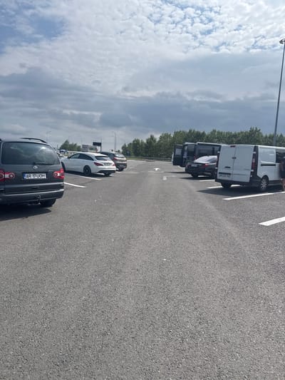A parking lot at a service area along a highway outside of Orăștie, Romania, features several vehicles. In the foreground, a dark gray Volkswagen Sharan minivan is parked on asphalt. White parking lines mark the spaces. To the right of the minivan, a white Mercedes-Benz CLA station wagon is parked, followed by a dark-colored sedan and a white van. Several other vehicles, including a dark van and a lighter van, are parked further back. The environment is an open, paved area with sparse trees and vegetation in the background. The sky is partly cloudy with a mix of white and gray clouds. Streetlights are visible along the edge of the parking lot.
