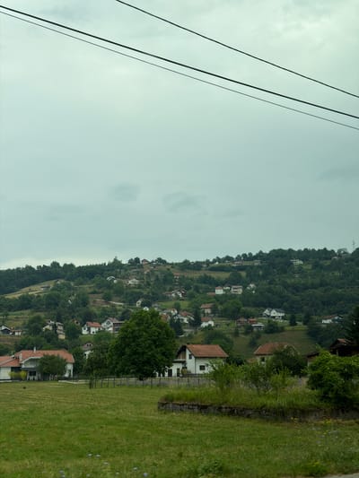 The image shows a hilly landscape with scattered houses and dense green trees under a cloudy sky. In the foreground, there is a large grassy field. Several houses, varying in size and architectural style, are visible on the slopes of the hills. Some houses have red tile roofs, while others have darker roofing. The vegetation is lush, with a mix of deciduous and coniferous trees. The overall impression is of a rural or semi-rural setting. The scene is likely located in Rastovci, Bosnia and Herzegovina, given the context provided. There are no people or animals visible in the image. The image is taken from a slightly elevated perspective. Power lines are visible in the upper portion of the image.