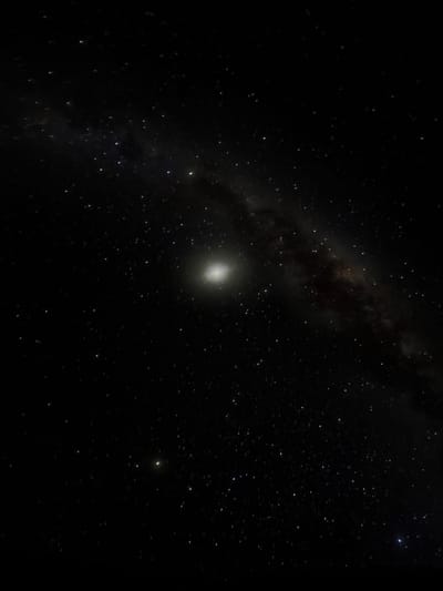 The image is a vertical astrophotograph of the night sky, captured from a location near Canela, Brazil. It features a prominent, bright elliptical galaxy with a concentrated luminous core, positioned slightly above the vertical center. Numerous individual stars of varying magnitudes are scattered across the dark expanse. Faint, diffuse dark lanes, consistent with interstellar dust or nebulosity, extend diagonally from the upper right to the lower left. A single, less luminous star is visible in the lower left quadrant. The environment depicted is deep space, observed under clear night conditions, devoid of terrestrial elements, people, or artificial light sources.