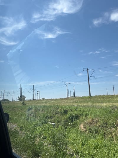 This is a daytime, outdoor photograph taken from a vehicle, looking out over a grassy embankment towards a clear blue sky with scattered white clouds. The foreground features the edge of the vehicle's window and a dark interior mirror. The midground is dominated by a lush, green, overgrown grassy field. Beyond the field, a series of tall, dark utility poles with overhead wires, characteristic of a railway line, stretch across the landscape. In the distance, there is a small building with a peaked roof, likely a signal box or small station building, positioned between the utility poles. The overall scene suggests a rural or suburban area with active railway infrastructure.