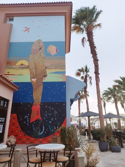 The image shows a view of a beachfront restaurant or cafe in Ras Abu Soma, Egypt. A mural adorns the side of a peach-colored building, showing a person-like figure from head-to-toe over various background scapes, including birds in the sky, a sunset over water, a large body of water, and plants in a black space. Tables and chairs, as well as large potted plants, are situated on a wooden deck in the foreground. Beyond the deck, the restaurant area extends towards the beach, with palm trees and several umbrellas. People can be seen on the beach in the distance.