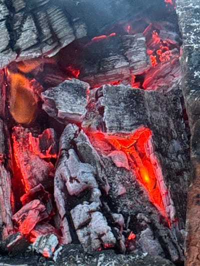 The image shows a close-up view of glowing embers and charred wood. The texture of the wood is rough and broken, with some pieces appearing to be partially consumed by fire. The embers glow with a bright red and orange light, illuminating the charred surfaces. Wisps of smoke are visible in the upper part of the image. The scene is dark and intense, with the fire being the primary source of light and color. There are no people or animals visible in the image. The image does not contain any contextual information related to the city of Imelevo, Russia, nor does it depict any specific location within a city. It is a generic representation of burning wood.