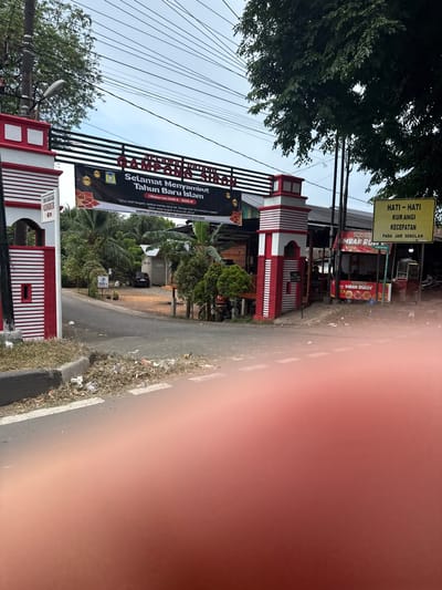 The image shows an outdoor scene in the city of Siron, Indonesia, with an arched gateway in the foreground. The gateway is red and white and has a banner that reads "SELAMAT DATANG GAMPONG SIREN" (Welcome to Gampong Siren) and "Selamat Menyambut Tahun Baru Islam" (Happy Islamic New Year). There are trees and buildings in the background. A yellow sign on the right reads "HATI-HATI KURANGI KECEPATAN PADA JAM SEKOLAH" (Be careful, reduce speed during school hours). Trash is visible on the side of the road. The sky is overcast.