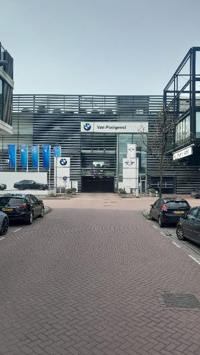 This image captures the exterior of a BMW Van Poelgeest car dealership in Amsterdam-Duivendrecht, Netherlands, under an overcast sky. The modern, multi-story building features a facade composed of dark horizontal slats and large glass panels, with a central recessed glass entrance. A large BMW logo and "Van Poelgeest" text are displayed above the main entrance. To the left of the entrance, several blue vertical banners with BMW branding, including "BMW Premium Selection" text, are visible alongside a vertical sign featuring the BMW logo. To the right of the entrance, a vertical sign displays the MINI logo. The foreground consists of a wide reddish-brown brick-paved road, laid in a herringbone pattern, with a square manhole cover visible in the middle-right. Parked vehicles line both sides of the road: on the left, a dark SUV (Dutch license plate 24-GZ-SS) and partially visible dark sedan are parked diagonally. On the right, a dark gray hatchback (Dutch license plate L-197-XP) and another dark small car are parked parallel to a concrete curb. Elevated display platforms to the left of the building showcase at least one dark sedan. A bare-branched tree stands to the right of the main entrance. No individuals are visible in the scene.