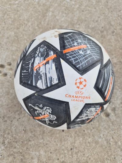 This media file features a close-up, top-down view of a UEFA Champions League soccer ball resting on a textured, concrete surface. The ball itself is predominantly white with black and orange paneling, adorned with the UEFA Champions League logo and various graphics. The design includes stylized imagery, potentially representing iconic cityscapes or stadiums associated with past finals, and a depiction of a lion. Text is visible on the ball, including "UEFA CHAMPIONS LEAGUE," and fragments of other text such as "2019 MADRID" and "2002 GLASGOW" can be discerned on some of the panels. The concrete surface is a light gray with a rough, uneven texture, showing some dirt and small indentations. No people are visible in the frame, and the lighting suggests it is daytime, with no specific indicators of weather conditions. The setting appears to be an outdoor, urban environment, given the concrete and the nature of the object.