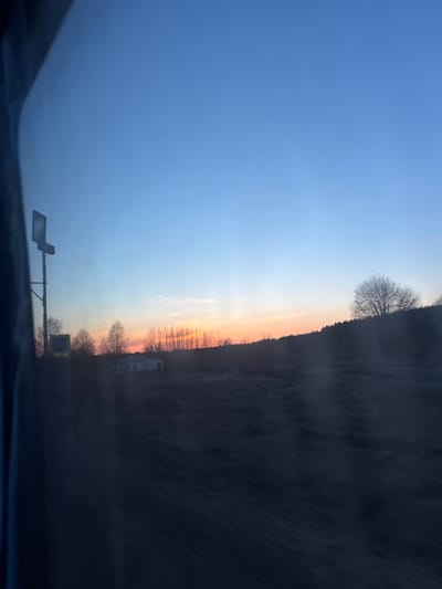 The image shows a view from inside a vehicle, likely a car, during sunrise or sunset. The sky transitions from a light blue to an orange-yellow horizon. In the distance, a treeline and what appears to be a building (possibly residential or commercial) are visible. There is also a traffic sign on the left side of the road. The perspective suggests the photo was taken while the vehicle was in motion. The location in Krasnye Gorki, Russia is not definitively ascertainable based on the visual information alone.