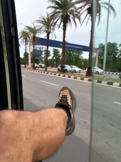 The image, taken from inside a vehicle, shows a person's leg and foot sticking out of a window. The foot is clad in a brown and black athletic shoe. The leg is hairy. Outside the vehicle, a road is visible with several cars driving on it. Palm trees line the road, and a blue billboard is present in the background. The sky is overcast. The location is identified as Gollapalle Khurd, India.