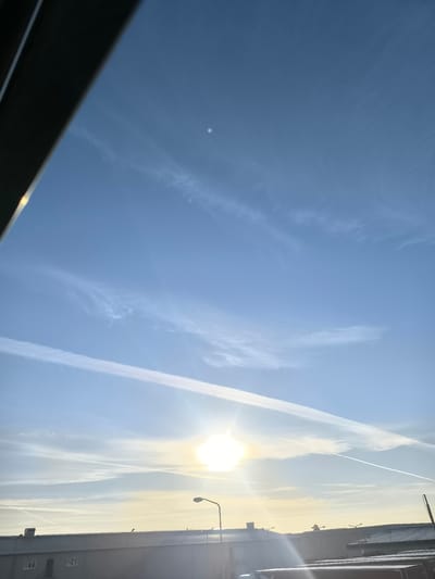 This image captures a bright, sunny sky over Uithoorn, Netherlands. The sun is prominently featured, casting a bright glare. Several contrails from airplanes streak across the clear blue sky, some appearing wispy and others more defined. In the lower portion of the image, the rooftops of industrial buildings are visible, with a streetlamp and other structures suggesting an urban or semi-industrial environment. The overall impression is one of a clear day with ongoing air traffic above the Dutch landscape.