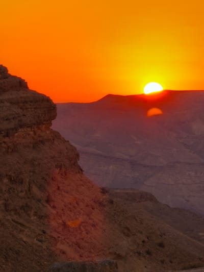 This is a vertical, medium shot of a desert landscape at sunset. The foreground features a steep, rocky cliff face on the left side of the image, illuminated by the setting sun. The cliff is textured with various shades of brown and red. In the background, rolling desert hills stretch towards the horizon. The sky is a vibrant gradient of orange and yellow, with the sun partially visible just above the distant hills. A faint, distorted reflection of the sun is visible on the hills in the middle ground. The scene is devoid of any people or man-made structures, suggesting a natural, remote setting. This type of dramatic sunset over the desert is characteristic of the Ramon Crater area near Yeruham, Israel, known for its unique geological formations and breathtaking natural beauty.