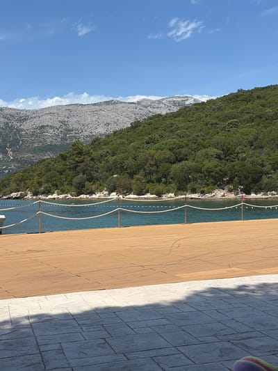 The image captures a scenic coastal view in Grad Korčula, Croatia, under a clear blue sky with sparse white clouds. In the background, a large, rugged grey mountain range with visible rock formations and minimal vegetation dominates the upper left, transitioning into a dense, green forested hill that slopes down towards the sea on the right. Below the forested hill, a light-colored, rocky shoreline marks the edge of a calm, turquoise-blue body of water. Several small human figures are discernible on the distant shoreline, suggesting bathers or individuals near the water's edge, with one person in a bright top visible towards the right side of the water. A safety rope barrier, consisting of two white ropes strung between six slender metal posts, extends horizontally across the mid-ground, delineating an area of the water and separating it from a light brown wooden deck. Beyond this rope, small white buoyant markers are scattered across the water surface. The foreground consists of this expansive wooden deck, which exhibits subtle scuff marks or tire tracks on its surface. Directly below and in front of the wooden deck is a grey, patterned stone-paved patio, cut by a sharp shadow line that covers approximately half of its surface. A small, multi-colored object with purple, green, and yellow stripes is partially visible in the bottom right corner.
