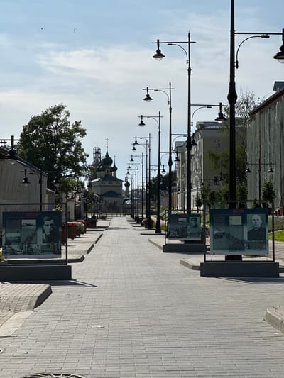 The image depicts a cobblestone street in the city of Nikul'skoe (Neverkovskii S/O), Russia, on a clear, sunny day. A row of traditional streetlights lines the left side of the street, with their ornate black metal poles and globes casting shadows on the pavement. On the right side of the street, buildings with light-colored facades are visible, some with scaffolding.
Several large display boards are strategically placed along the sidewalk, featuring portraits and historical information, likely related to local heroes or significant events. These boards are framed in dark metal and stand on individual bases. In the distance, a prominent church with multiple onion domes and a cross on top dominates the skyline, suggesting this is a historically significant area. Trees and planters filled with flowers add touches of greenery and color to the urban landscape. The sky above is a pale blue with scattered white clouds, and two birds are visible in flight. The street appears quiet, with no people in the immediate foreground.