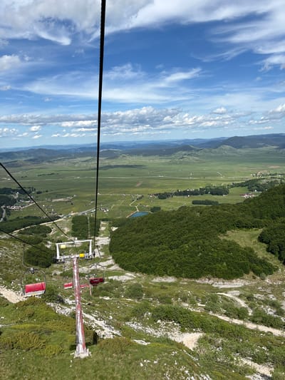 An aerial shot captures a scenic view of Pašina Voda, Montenegro, from a ski lift. The ski lift, with its red and white pole and chairs, is prominently featured in the foreground. Below, a lush green valley unfolds, dotted with small houses and a tranquil blue lake. Rolling hills and mountains form the backdrop, under a sky filled with dramatic white clouds. The landscape is a blend of natural beauty and subtle human presence.