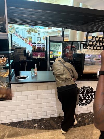 The photo shows an indoor scene, likely a coffee shop franchise in a mall, with customers at the counter. One customer, wearing a khaki jacket and black pants, is interacting with the shop employee standing behind the counter. The coffee shop is identified by the "SOUTH AFRICA'S PRESSO" logo on the wall. The menu is displayed, including a list of prices for juices and slushies. The image also contains a clear depiction of the interior decor, showing a detailed view of the surroundings. The location is not explicitly mentioned in the image itself, however the OCR mentions the location as Milnerton, South Africa.