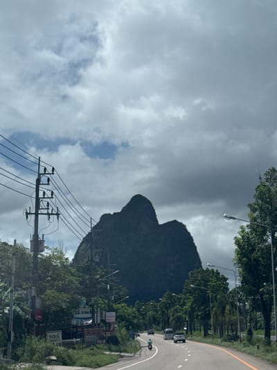 This media file depicts a road running alongside a lush, green landscape dominated by a large, dark, mountainous rock formation. The sky is heavily overcast with dramatic, grey clouds, suggesting a gloomy or possibly rainy day. A winding road, marked with white lines and a yellow line on the right edge, is visible, with a few cars on it. Tall trees and utility poles with numerous wires line the roadside, creating a sense of a rural or semi-urban environment. The rock formation itself is imposing, with a distinct, almost heart-like shape visible in the center. The overall impression is one of a natural landscape being traversed by modern infrastructure. There is some visible text on signs, but it is too blurry to read clearly. The time of day appears to be daytime, given the ambient light. The weather conditions are clearly cloudy and potentially humid, fitting the tropical setting of Phang-nga, Thailand.