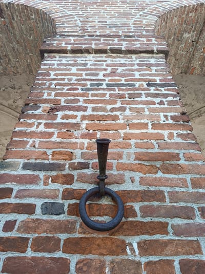A low-angle, full shot depicts a brick wall with a metal ring and holder affixed to it. The wall is constructed of red and brown bricks with white mortar, and it forms part of an archway. The metal ring is black and has a holder that extends upwards from it. The wall is weathered and shows signs of age. The archway is situated in Mezzana Sant'Antonio, Italy.