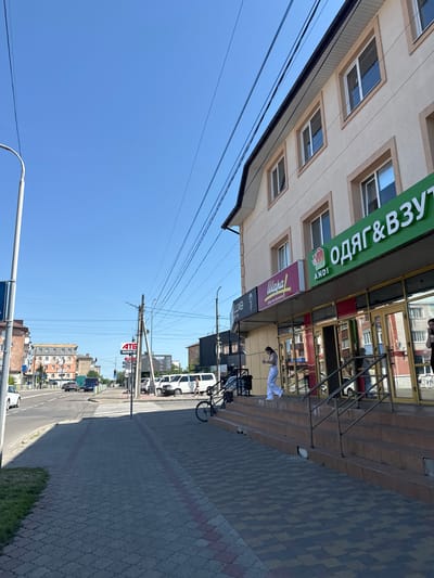 The image captures a street scene in the city of Sarny, Ukraine, on a clear, sunny day. In the foreground, a paved sidewalk with a decorative pattern runs alongside a multi-story building. The building's facade is light-colored, with several windows and signage. Prominent green signage with white lettering reads "ОДЯГ&ВЗУТ" (Clothing & Shoes), along with "АНДІ" (ANDI). Below this, other signs are visible, including one with a stylized flower and "Luxe" in smaller lettering, and another with a red and white design and "Шаг" (Step).
The building has large glass-fronted shop entrances with metal railings leading up a set of concrete stairs. A young woman in white pants and a white top is walking up these stairs, holding a cup. A bicycle is parked to the right of the entrance.
Beyond the building, a street is visible with several vehicles, including cars and vans. A pedestrian crossing is marked on the road. On the left side of the street, more buildings and a traffic light can be seen. Overhead, power lines crisscross the sky, and a tall pole with a street light stands on the left side of the sidewalk. The sky is a vibrant blue.