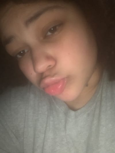 The image depicts a close-up portrait of an individual, likely female, from the chest up, positioned against a dark, indistinct background. The subject has medium-toned skin, dark eyes, and dark hair, part of which is visible on the right side of the frame. The individual is wearing a grey, crew-neck t-shirt. Their head is tilted slightly. The eyes are open and directed towards the viewer. The most prominent feature is the mouth, with lips pursed forward in a "duck face" pose, appearing glossy and reddish-pink. There are no other discernible objects or subjects within the frame. The lighting is low, illuminating the face from the front. The environment provides no identifying details of a specific location within the city of Lancaster, United States of America, appearing to be an indoor setting with minimal background context.