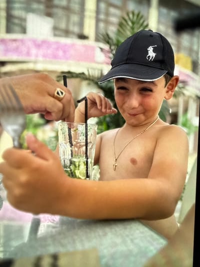 A young boy wearing a black baseball cap with a white polo logo and a gold cross necklace is seen from the chest up. He has a mischievous expression, with his mouth slightly open and eyes squinted. His skin is flushed, suggesting he might be warm or excited. Two hands are visible in the foreground, one on the left holding a fork and another on the right stirring a drink in a glass with two black straws. The drink appears to be a clear liquid with green slices, possibly lime. The background is blurred but suggests an outdoor setting with pink and white structures and greenery. The image appears to be taken in an outdoor restaurant or cafe, possibly in Artik, Armenia, judging by the context.
