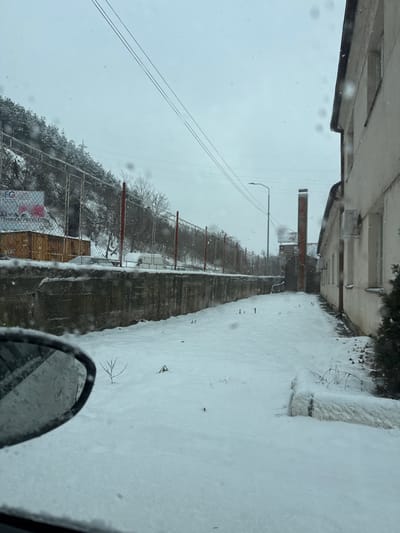 The image displays an outdoor scene during an overcast winter day with ongoing light snowfall, as indicated by visible flakes and a uniform layer of snow covering all horizontal surfaces. The vantage point is from inside a vehicle, evidenced by the upper portion of a side mirror in the lower-left foreground and moisture on the window.

The scene is located in the city of Raska, Raska Municipality, Serbia.

On the left, a steep, snow-covered wooded hillside rises, secured by a chain-link fence with prominent red vertical posts. Below the fence runs a substantial, weathered concrete retaining wall or platform. Adjacent to this wall, a wooden hoarding structure exhibits a partially visible sign bearing "FG" and "TEHNIČKI PREGLED," along with a graphic of a red truck. Behind the concrete structure and fence, several snow-dusted vehicles, including a white van, are parked.

On the right, a two-story, cream-colored building with multiple windows and an external air conditioning unit runs parallel to the scene's progression.

The ground between the concrete wall and the building is an open, snow-covered expanse with sparse, emergent vegetation. In the midground, a tall, slender red-brick chimney is visible, suggesting industrial or commercial activity, alongside a street light pole. Multiple overhead power lines traverse the pale, cloud-dense sky. No people or active interactions are present in the frame; the scene is static under snowy conditions.