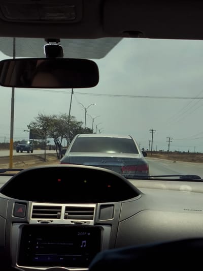 The image is taken from the interior of a car, focusing on the dashboard and the view through the windshield. The car appears to be driving on a paved road, possibly a highway, in the direction away from La Cruz del Pastel, Venezuela, as evidenced by the dry, arid landscape and the electrical poles visible in the distance. Another car is directly in front, and a dark-colored SUV is visible in the left lane. The car's infotainment system is displaying music information, indicating the song "Volvi" by Aventura and Bad Bunny is playing. The time displayed is 2:05. The overall scene suggests a routine daytime drive.