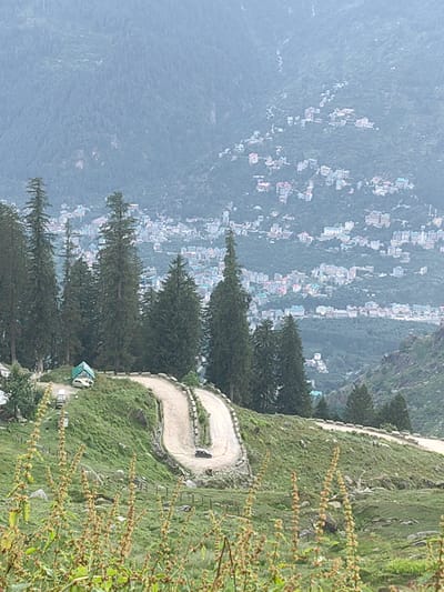 The image shows a scenic view of a winding road on a grassy hillside. In the foreground, there are tall, green trees and some dry, wispy plants. The road itself is made of a light-colored, dusty material and curves sharply up the hill. There is a single black car visible on the road. A small green structure, possibly a tent or a shed, is situated near the top of the hill, with a white car parked next to it. In the background, a town or city is spread across the slopes of a large, forested mountain. The buildings are varied in color, including white, pink, and teal, and appear to be clustered together. The overall environment is mountainous and somewhat hazy, suggesting a distance view or possibly fog. The location is identified as Chhyal, India.