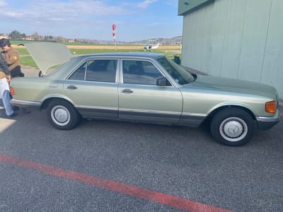 The scene depicts an outdoor environment at the Aerodrome de Chonas-l'Amballan, France, during daylight hours. A four-door, pale metallic green Mercedes-Benz W126 series sedan is centrally positioned, oriented towards the right of the frame. The vehicle's trunk lid is open. Two individuals are partially visible at the rear-left of the car; one is wearing a brown jacket with a circular emblem on the left arm, and dark pants, while the other is wearing light blue jeans and dark shoes. They appear to be standing near the open trunk. The ground is dark asphalt tarmac, featuring a faded, broad red line painted horizontally in the foreground. To the right, a large light green corrugated metal hangar wall is visible. In the background, a grassy airfield extends, featuring a prominent white and red striped windsock and a small white propeller aircraft with dark markings. Distant rolling hills with scattered deciduous trees are visible under a light blue sky with scattered white clouds.