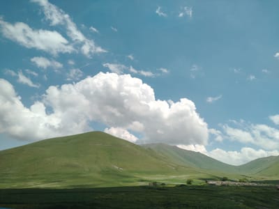 The image depicts a vast, rolling green landscape under a bright blue sky dotted with fluffy white clouds. In the foreground, a dark green grassy field stretches across the bottom of the frame. Behind this, a large, verdant hill dominates the left side of the image, its slopes curving gently upwards. Further back, a series of progressively receding hills and mountains form a valley. On the right side of the midground, a long, low-rise building with a light-colored roof and a fence in front of it is visible, with a few trees scattered around it. The overall impression is one of natural beauty and tranquility. This scene is located in Jrashen, Armenia, characterized by its expansive green pastures and mountainous terrain.