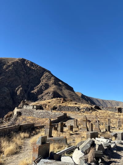 A rugged mountain range meets a clear blue sky. On a steep hillside, an ancient cemetery with weathered stone structures and crosses stands amidst dry grass. Retaining walls are constructed along the slope. The scene evokes a sense of historical significance and remote isolation under bright sunlight.