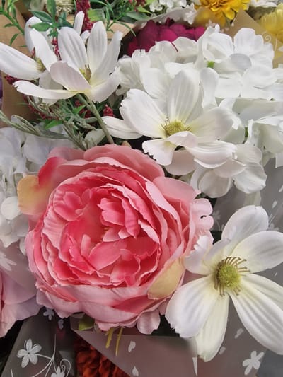 This media file is a close-up, eye-level shot of a bouquet of artificial flowers. The arrangement features a prominent, large pink peony in the foreground, with its layered petals in various shades of pink. Behind it, several white flowers with delicate, elongated petals and green centers are visible, appearing to be a type of magnolia or starflower. There are also hints of other flowers, including yellow blooms and a deep magenta flower. The bouquet is presented against a backdrop that includes some decorative wrapping paper with a subtle floral pattern and a plain brown surface, possibly a table or part of the arrangement's packaging. The lighting is soft and diffused, suggesting an indoor setting or an overcast day. There are no people or specific time-of-day indicators. The overall impression is one of decorative floral display, likely intended for an event or as a gift. The image does not contain any visible text.
