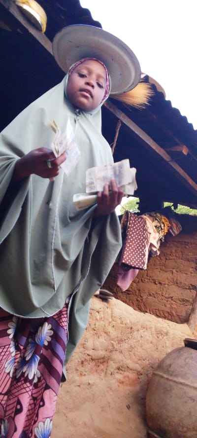 In Yayu, Nigeria, a young girl wearing a light green hijab and a patterned skirt stands outdoors. She is holding several banknotes in her hands, appearing to be displaying them. A large metal tray is balanced on her head, and a broom is visible behind her. The background consists of a rustic structure with a corrugated metal roof and mud walls, where various colorful cloths are hung. The ground is made of earth and dirt, and a large metal pot is situated to the right. The scene suggests a market or roadside stall setting.