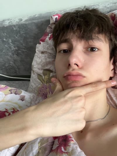 The scene is an interior view, likely a bedroom, situated in the city of Zainsk, Russia. A young individual with fair skin, dark brown short hair, and brown eyes is depicted in a close-up, head-and-shoulders portrait, lying on their back. The person's lips are slightly pursed, forming a pout. Their right hand is raised, with the index finger extended and touching the underside of their chin, while the thumb rests near the jawline. A thin, silver-colored necklace is visible around their neck. The individual is partially covered by bedding, which includes a pillow and blanket featuring a floral pattern with red, pink, purple, and green elements on a light background. In the upper left background, behind the pillow, a plain dark grey textured surface, possibly a headboard or wall, is visible. The lighting is even, suggesting an indoor setting.
