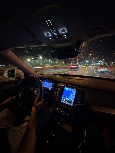 The image shows the interior of a car at night, positioned from the driver's perspective. The car appears to be stationary, with a view of a brightly lit street in front. Several cars and streetlights are visible. The car's dashboard and infotainment system are illuminated, displaying navigation information. The driver's hands are on the steering wheel. The interior lights of the car are partially lit. The location is presumed to be in Zeytinburnu, Turkey.