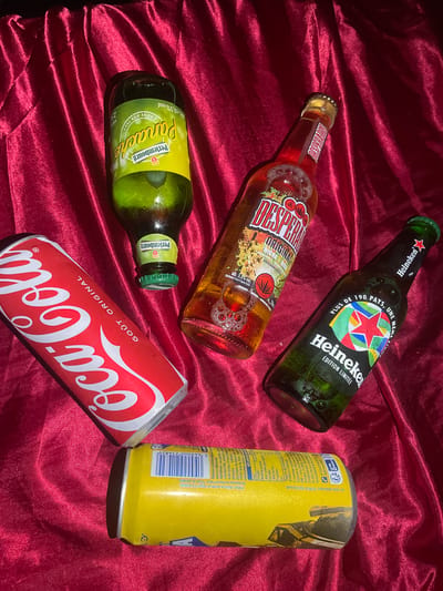 The image shows a collection of beverages arranged on a red, draped surface. The beverages include a bottle of Perrier, a bottle of Desperados, a bottle of Heineken, a can of Coca-Cola, and a yellow can with French text. The Perrier bottle has a green label with yellow text, and the neck is covered by a green cap. The Desperados bottle is filled with an amber liquid and has a label with red, white, and yellow lettering. The Heineken bottle is green and features a label with a red star and multi-colored circles, along with the Heineken logo. The Coca-Cola can is red with white lettering. The yellow can has blue and white text and graphics. The red fabric appears to be a tablecloth or draped material, with visible folds and creases. There is no indication of the city of Gerstheim, France, or any specific location within it. The scene is static and there are no people or actions depicted.