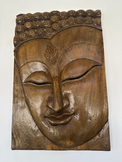 A wooden carving of Buddha's face dominates the image. The face, smooth and polished, features closed eyes, a prominent nose, and serene lips. A stylized lotus flower adorns the forehead. The left side of the carving showcases a series of tightly-wound circular patterns representing Buddha's hair. The wood grain is visible throughout, enhancing the artistic texture. The carving is set against a plain white wall.