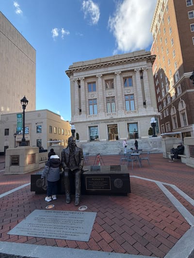 A statue of Joel Roberts Poinsett sits in Court Square, with a monument providing historical information. Behind the statue, steps lead up to M. Judson Booksellers and Camilla Kitchen, located at 130 Court Square, in Greenville, SC. A modern building is on the left, while a historical brick building looms in the background, against a blue sky. A child, wearing a hat and backpack, approaches the statue. The brick-paved square is partially enclosed with a light gray stone border.